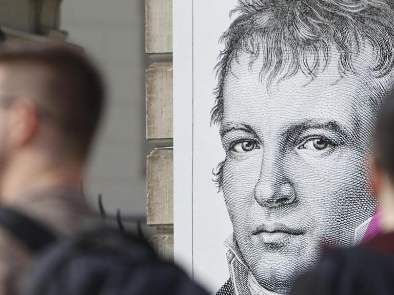 Menschen gehen an einer Plakatwand mit einem Bild des preußischen Forschers, Geographen und Naturforschers Alexander von Humboldt vorbei.