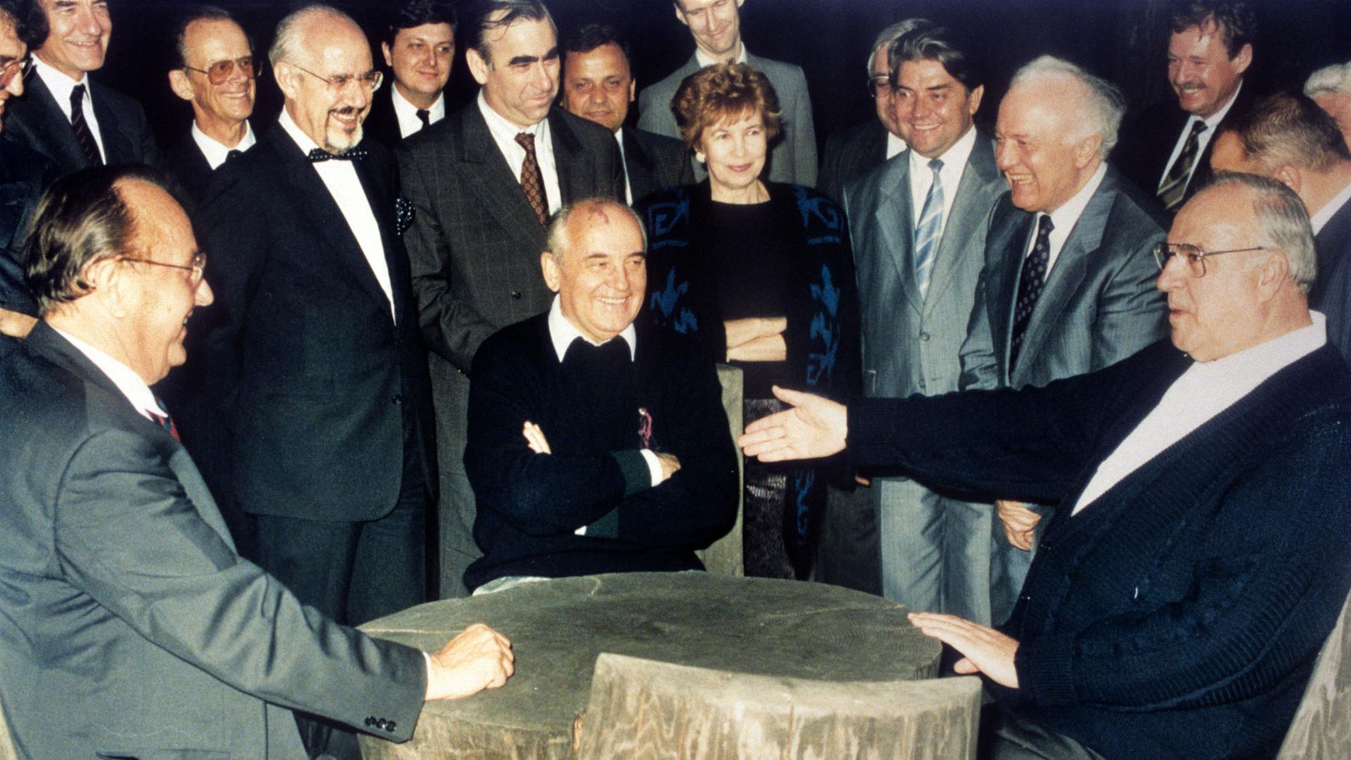 Bundeskanzler Helmut Kohl (r), der sowjetische Staatspräsident Michail Gorbatschow (M) und Bundesaußenminister Hans-Dietrich Genscher (l) unterhalten sich am 15.07.1990 in entspannter Atmosphäre an einem rustikalen Arbeitstisch in der freien russischen Natur, während die anderen Gäste unter anderem Raissa Gorbatschowa (M hinten) und Eduard Schewardnadse amüsiert die Szene betrachten. Bundeskanzler Helmut Kohl (r), der sowjetische Staatspräsident Michail Gorbatschow (M) und Bundesaußenminister Hans-Dietrich Genscher (l) unterhalten sich am 15.07.1990 in entspannter Atmosphäre an einem rustikalen Arbeitstisch in der freien russischen Natur, während die anderen Gäste unter anderem Raissa Gorbatschowa (M hinten) und Eduard Schewardnadse amüsiert die Szene betrachten.