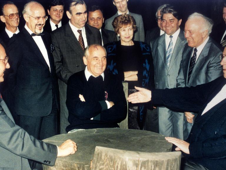 Bundeskanzler Helmut Kohl (r), der sowjetische Staatspräsident Michail Gorbatschow (M) und Bundesaußenminister Hans-Dietrich Genscher (l) unterhalten sich am 15.07.1990 in entspannter Atmosphäre an einem rustikalen Arbeitstisch in der freien russischen Natur, während die anderen Gäste unter anderem Raissa Gorbatschowa (M hinten) und Eduard Schewardnadse amüsiert die Szene betrachten.