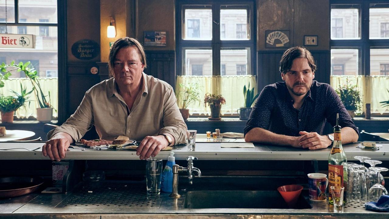 Peter Kurth und Daniel Brühl in einer Szene aus dem Film Nebenan. Sie sitzen nebeneinander an einer Theke in einer Bar.