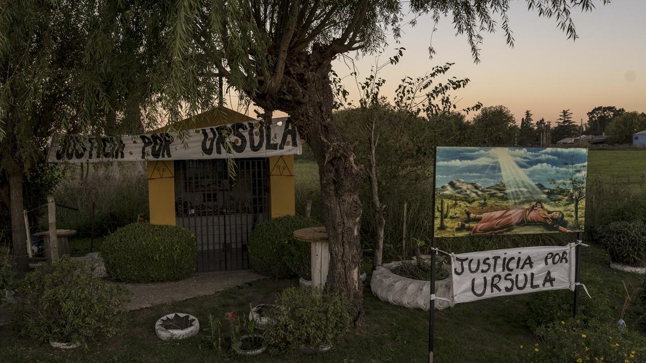 Eine Kapelle inmitten einer Grünanlage mit Bäumen und Büschen. Darüber zwei Banner, auf denen "Gerechtigkeit für Ursula" aus spanisch steht.