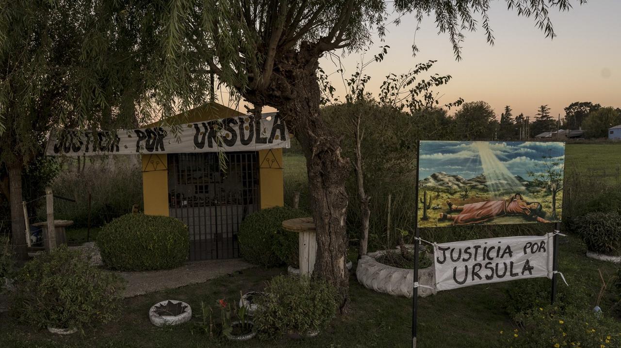 Eine Kapelle inmitten einer Grünanlage mit Bäumen und Büschen. Darüber zwei Banner, auf denen "Gerechtigkeit für Ursula" aus spanisch steht. Eine Kapelle inmitten einer Grünanlage mit Bäumen und Büschen. Darüber zwei Banner, auf denen "Gerechtigkeit für Ursula" aus spanisch steht.