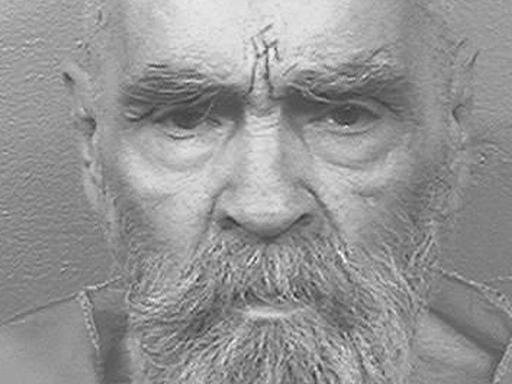 Charles Manson steht vor einer weißen Wand, auf der Stirn trägt er ein Hakenkreuz-Tattoo