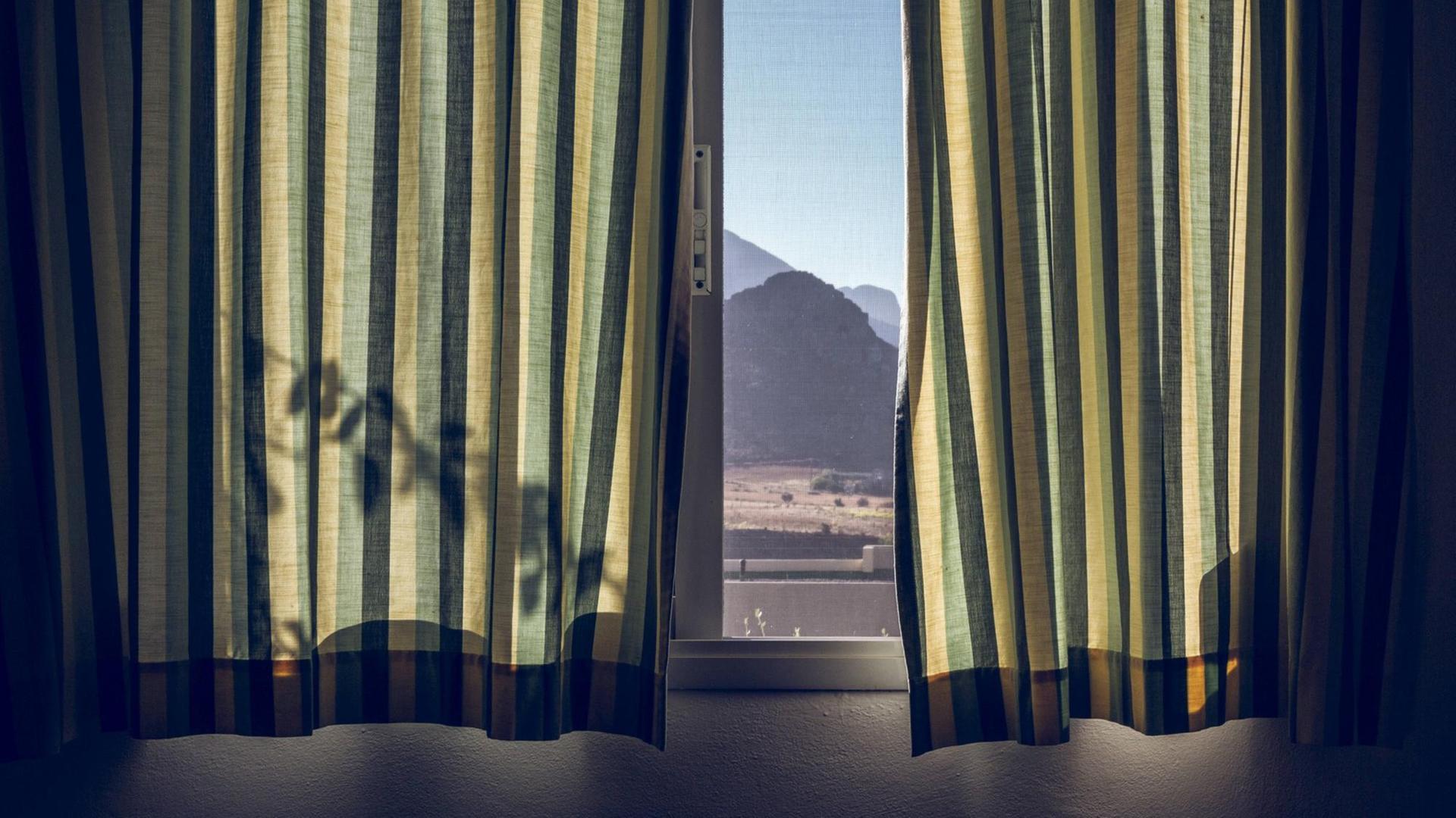 Durch ein Fenster mit fast ganz zugezogenen, grünen Vorhängen lässt sich eine bergige Landschaft erkennen. Durch ein Fenster mit fast ganz zugezogenen, grünen Vorhängen lässt sich eine bergige Landschaft erkennen.