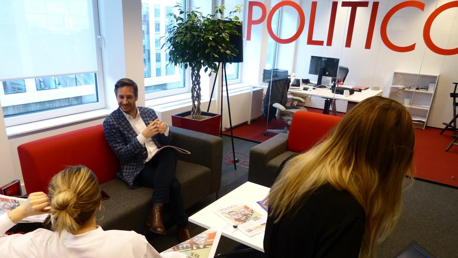 Drei Mitarbeiter von "Politico Europe" sitzen in den Brüsseler Redaktionsräumen, ein Schriftzug "Politico" ist auf einer Glaswand im Hintergrund zu lesen.