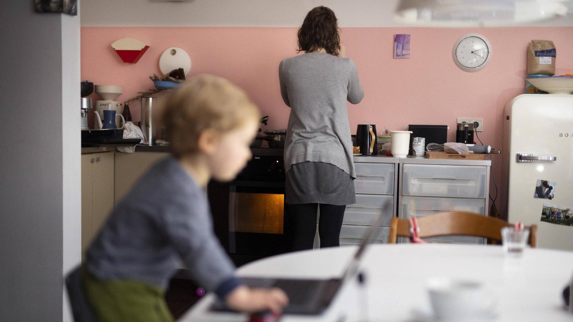Eine Frau steht mit dem Rücken zum Betrachter in einer modernen Küche am Herd. Im Vordergrund ist in der Unschäre ein kleiner Junge an einem Laptop zu sehen. Eine Frau steht mit dem Rücken zum Betrachter in einer modernen Küche am Herd. Im Vordergrund ist in der Unschäre ein kleiner Junge an einem Laptop zu sehen.