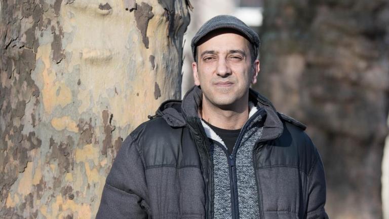 Porträt von Rouzbeh Taheri vor zwei Platanen. Er trägt eine gesteppte Daunenjacke und eine Mütze. Porträt von Rouzbeh Taheri vor zwei Platanen. Er trägt eine gesteppte Daunenjacke und eine Mütze.