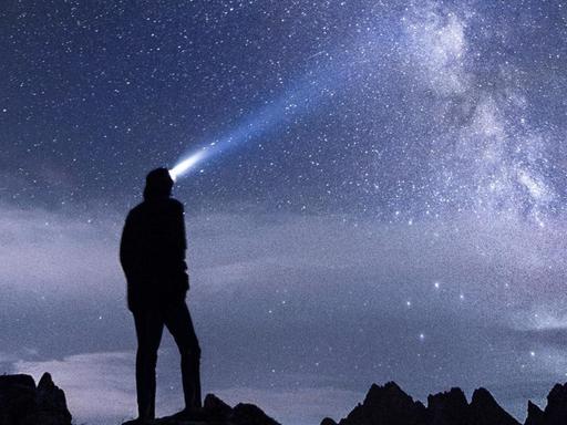 Silhouette eines Menschen der mit einer Strinlampe in den Sternenhimmel leuchtet.