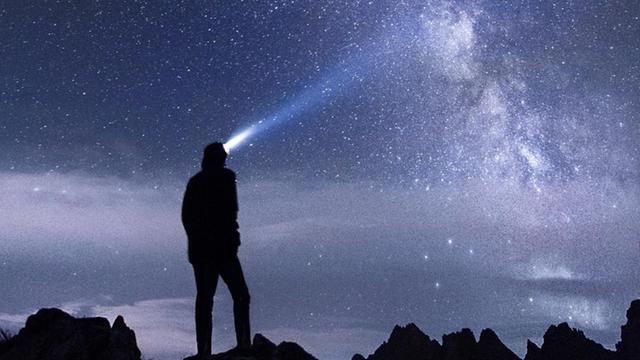 Silhouette eines Menschen der mit einer Strinlampe in den Sternenhimmel leuchtet.