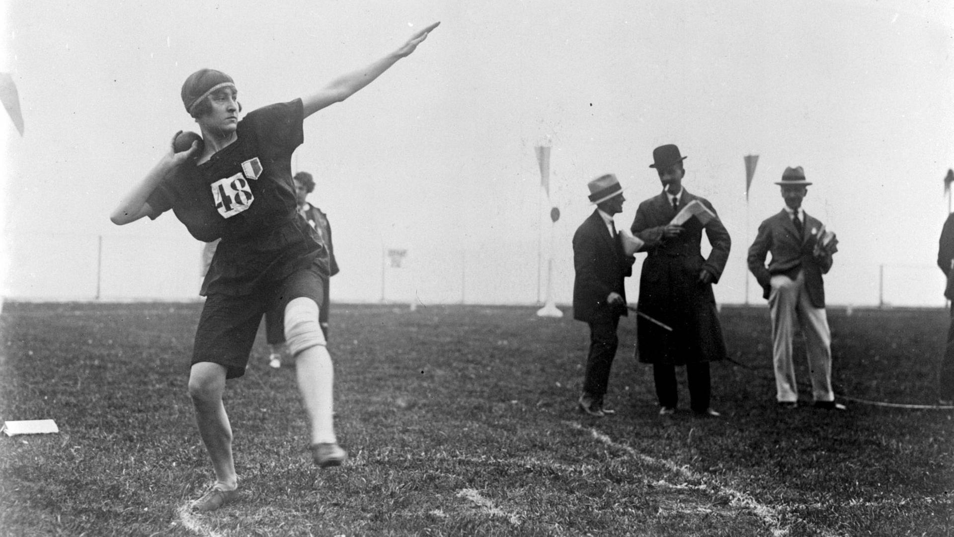 Ein Schwarzweiß-Foto zeigt eine Athletin mit einer Kappe, die mit einem Ausfallschritt Anlauf zum Kugelstoßen nimmt. Im Hintergrund sind zwei Männer mit Borsalino-Hüten ins Gespräch vertieft zu sehen