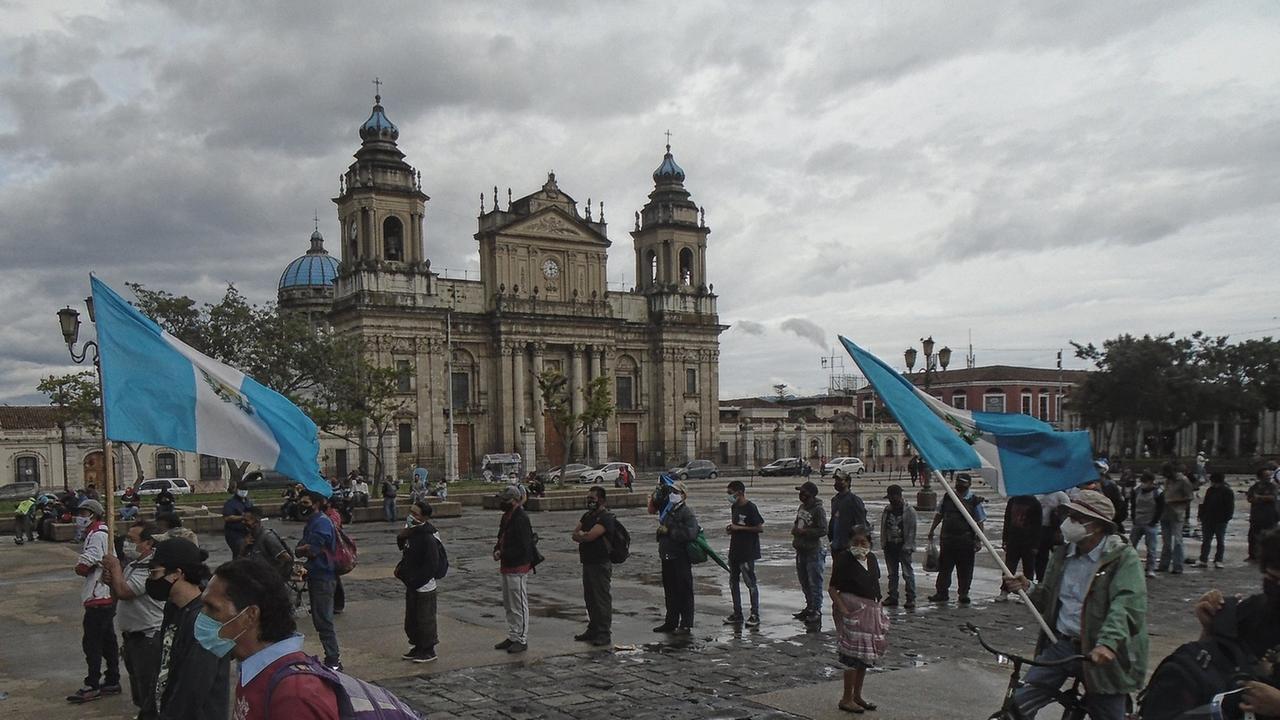 Demonstrierende stehen auf dem Platz vor der Kathedrale in Guatemala-Stadt, zwei von ihnen tragen blau-weiße Fahnen, alle tragen Mund-Nase-Masken und halten Abstand voneinander, um sich während der weltweiten Covid-19-Pandemie vor Ansteckung zu schützen, der Himmel ist wolkenverhangen. Demonstrierende stehen auf dem Platz vor der Kathedrale in Guatemala-Stadt, zwei von ihnen tragen blau-weiße Fahnen, alle tragen Mund-Nase-Masken und halten Abstand voneinander, um sich während der weltweiten Covid-19-Pandemie vor Ansteckung zu schützen, der Himmel ist wolkenverhangen.