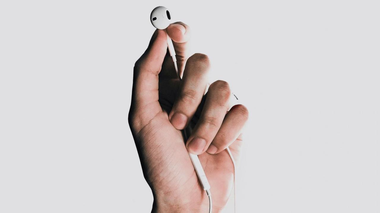 Fotografie einer Hand, die einen weißen In-Ear-Kopfhörer zwischen Daumen und Zeigenfinder hält. Fotografie einer Hand, die einen weißen In-Ear-Kopfhörer zwischen Daumen und Zeigenfinder hält.