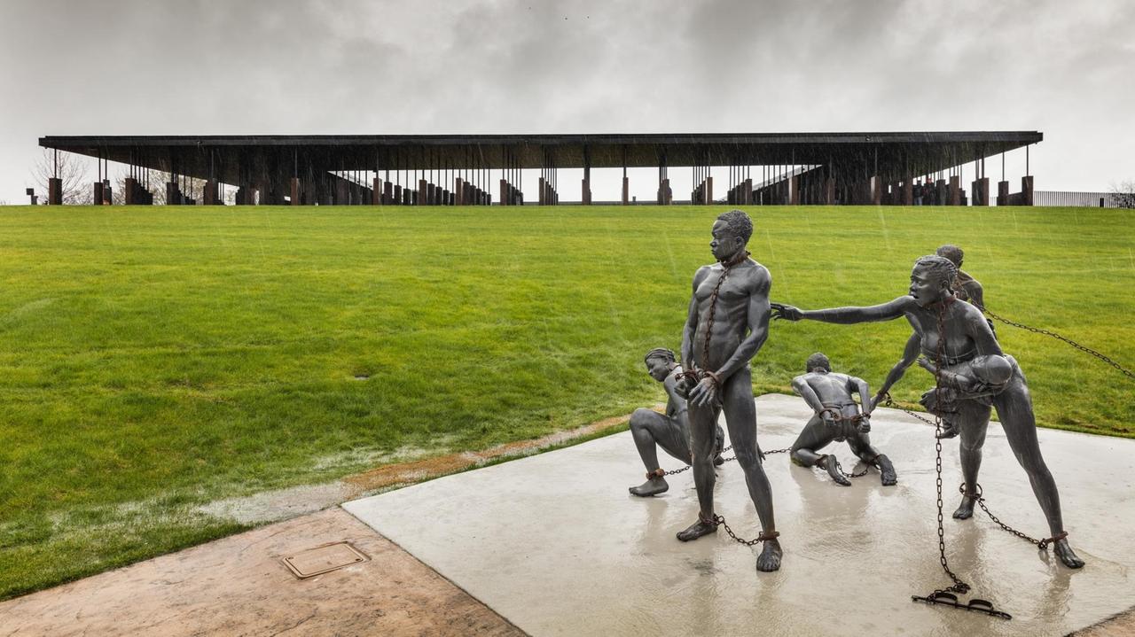 Die Skulpturengruppe "Nkyinkim" von Kwame Akoto-Bamfo ist den Opfern des transatlantischen Sklavenhandels gewidmet. Das "National Memorial for Peace and Justice" im Hintergrund ist eine nationale Gedenkstätte zur Erinnerung an die Opfer rassistischer Lynchjustiz in den Vereinigten Staaten.  (Montgomery, Alabama)