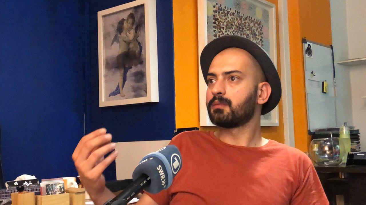 Maziyar Khavajian sitzt in seiner Wohnung mit Hut, schwarzem Vollbart und dunkelrotem T-Shirt vor dem Mikrofon und gestikuliert mit der rechten Hand. Maziyar Khavajian sitzt in seiner Wohnung mit Hut, schwarzem Vollbart und dunkelrotem T-Shirt vor dem Mikrofon und gestikuliert mit der rechten Hand.