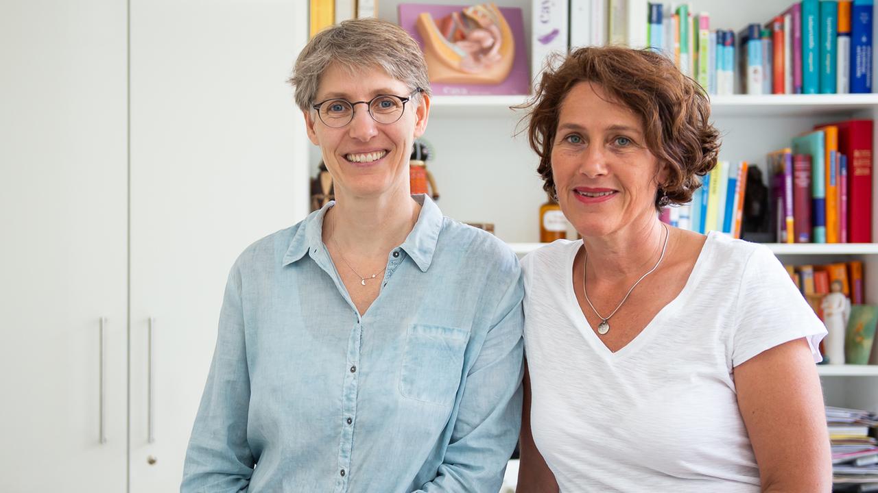 Die Frauenärztinnen Natascha Nicklaus (l.) und Nora Szász stehen vor einer Bücherwand und lächeln.