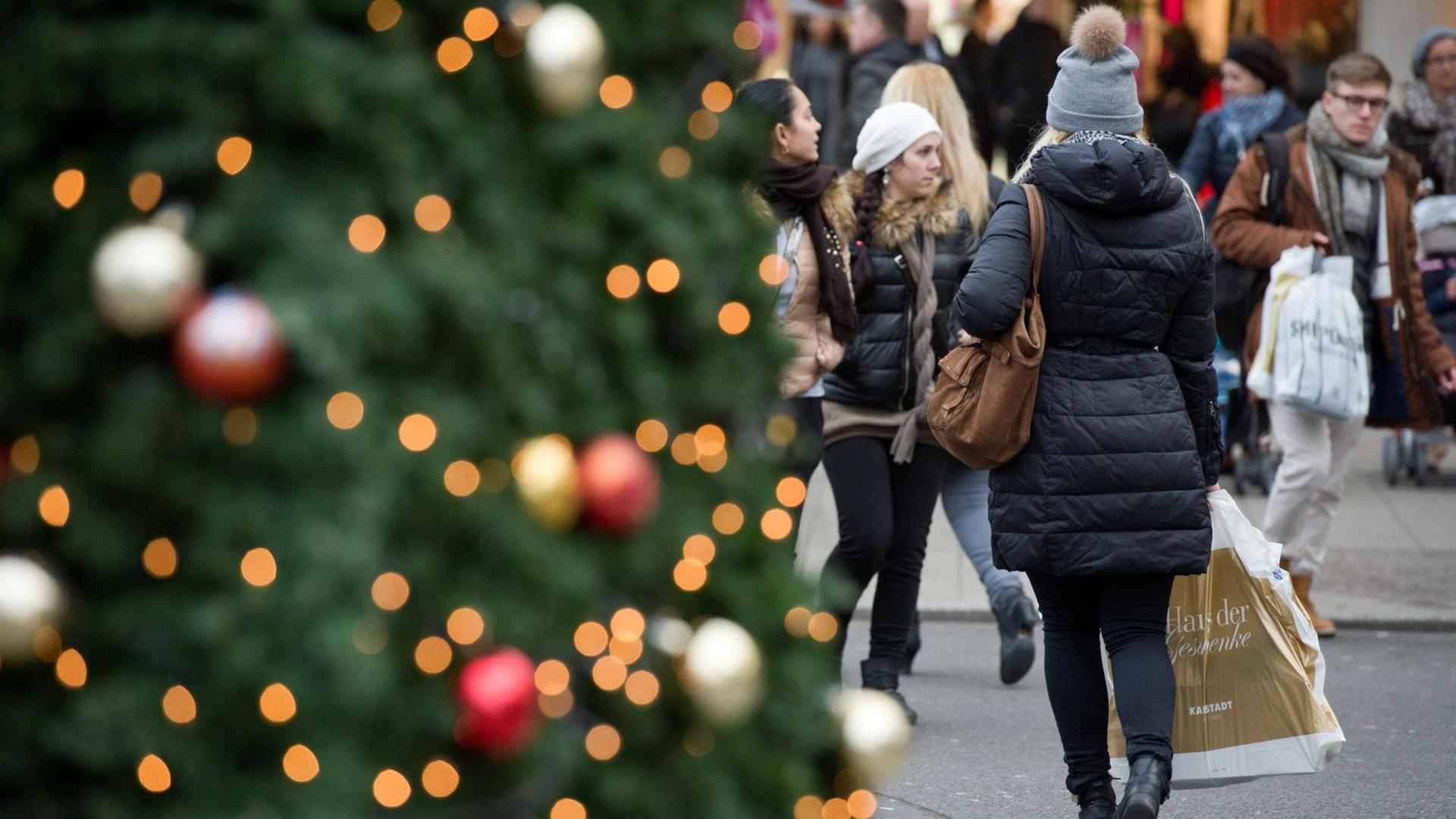 Links im Vordergrund ein Weihnachtsbaum, neben dem eine Frau mit einer Einkaufstüte entlang geht. Links im Vordergrund ein Weihnachtsbaum, neben dem eine Frau mit einer Einkaufstüte entlang geht.