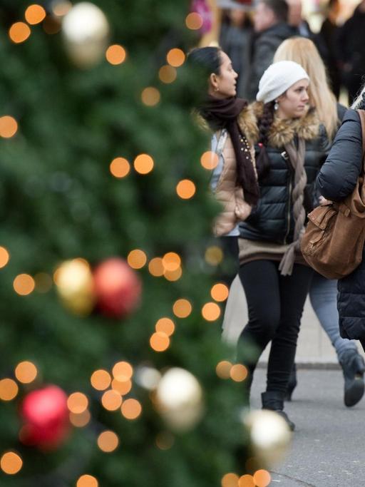 Links im Vordergrund ein Weihnachtsbaum, neben dem eine Frau mit einer Einkaufstüte entlang geht.