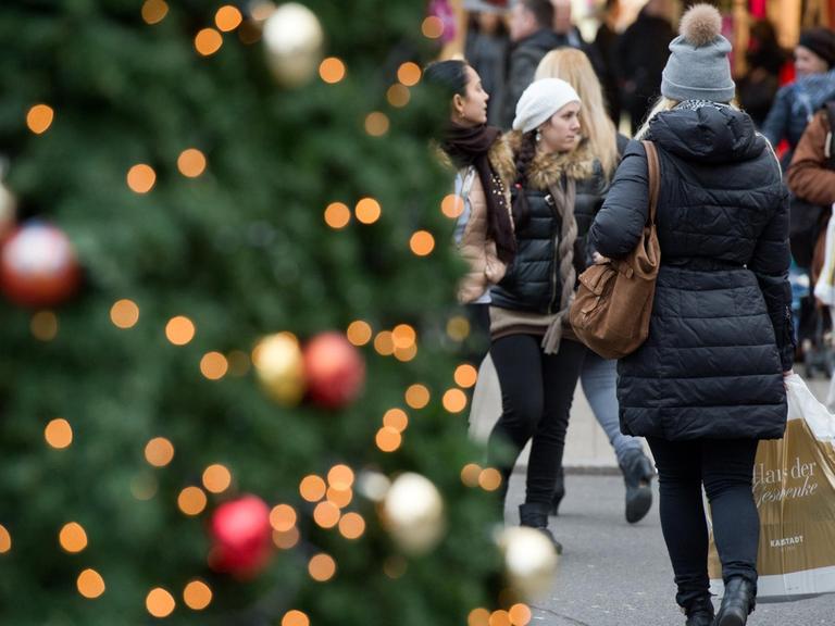 Links im Vordergrund ein Weihnachtsbaum, neben dem eine Frau mit einer Einkaufstüte entlang geht.