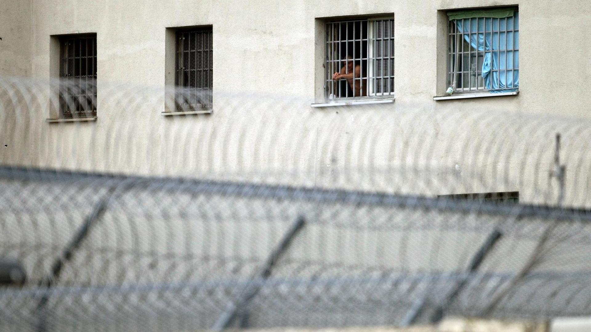Außenwand des Gefängnis in Leipzig mit vier vergitterten Fenstern, aus dem zweiten von rechts schau ein Mann. Davor befindet sich ein Sacheldrahtzaun