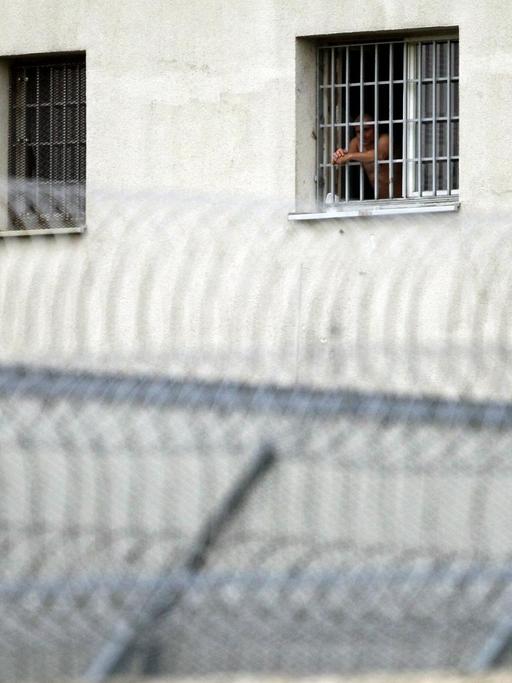 Außenwand des Gefängnis in Leipzig mit vier vergitterten Fenstern, aus dem zweiten von rechts schau ein Mann. Davor befindet sich ein Sacheldrahtzaun