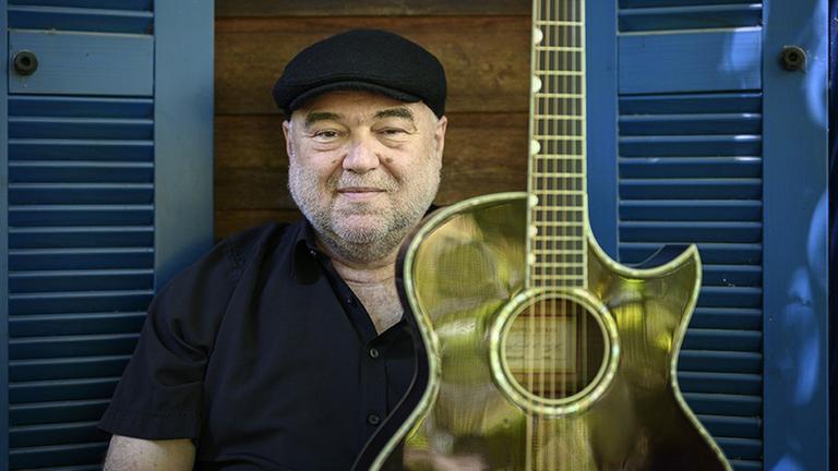 Ein Mann mit Kappe und einer Gitarre sitzt vor einem blauen geöffneten Fensterladen. Ein Mann mit Kappe und einer Gitarre sitzt vor einem blauen geöffneten Fensterladen.