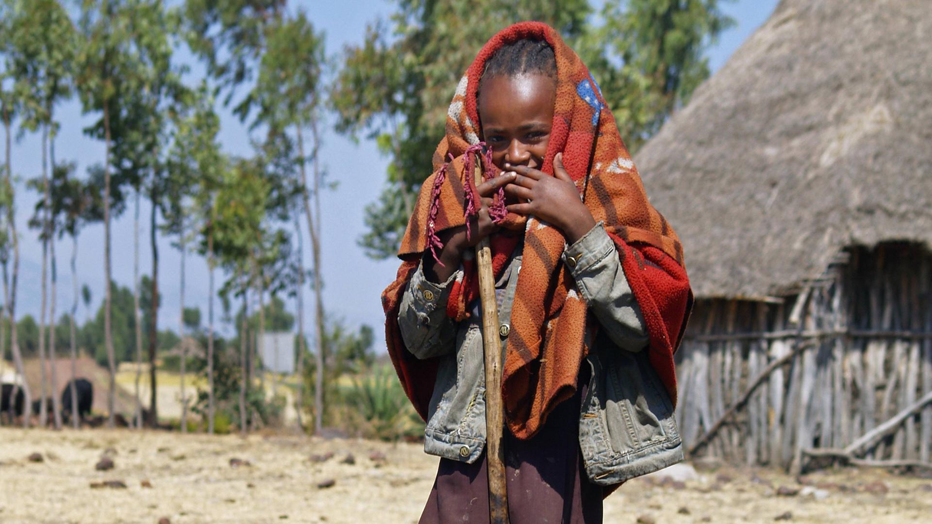 Mädchen mit Decke um den Kopf und Holzstock in der Hand vor einer Hütte in Uganda Mädchen mit Decke um den Kopf und Holzstock in der Hand vor einer Hütte in Uganda