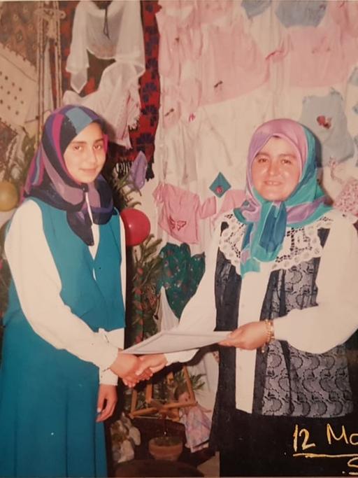 Foto eines analogen Fotos vom 12. Mai 1992: Eine ältere Frau gibt einer jüngeren die Hand und überreicht ihr ein Dokument. Beide tragen ein Kopftuch. Im Hintergrund ist jede Menge Kinderkleidung und Blumen zu sehen.