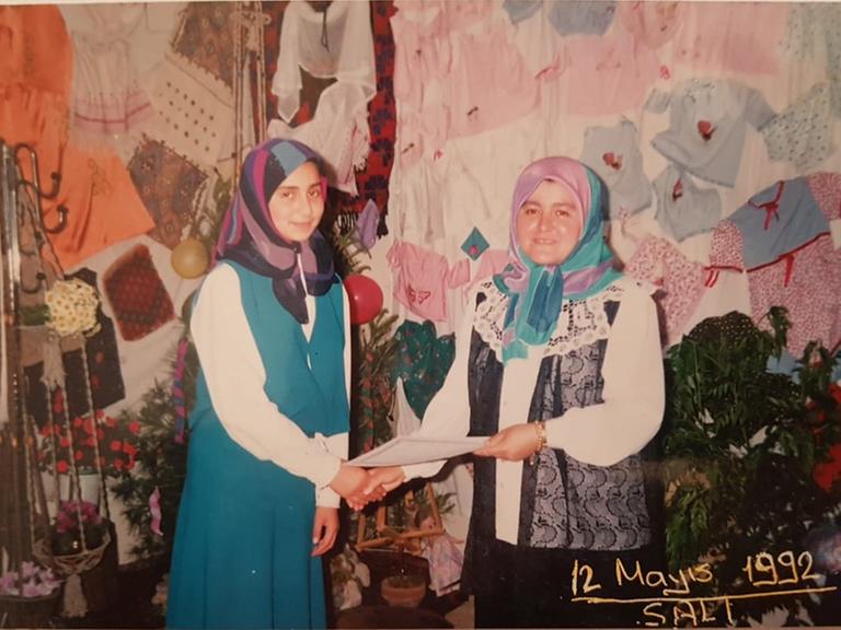 Foto eines analogen Fotos vom 12. Mai 1992: Eine ältere Frau gibt einer jüngeren die Hand und überreicht ihr ein Dokument. Beide tragen ein Kopftuch. Im Hintergrund ist jede Menge Kinderkleidung und Blumen zu sehen.