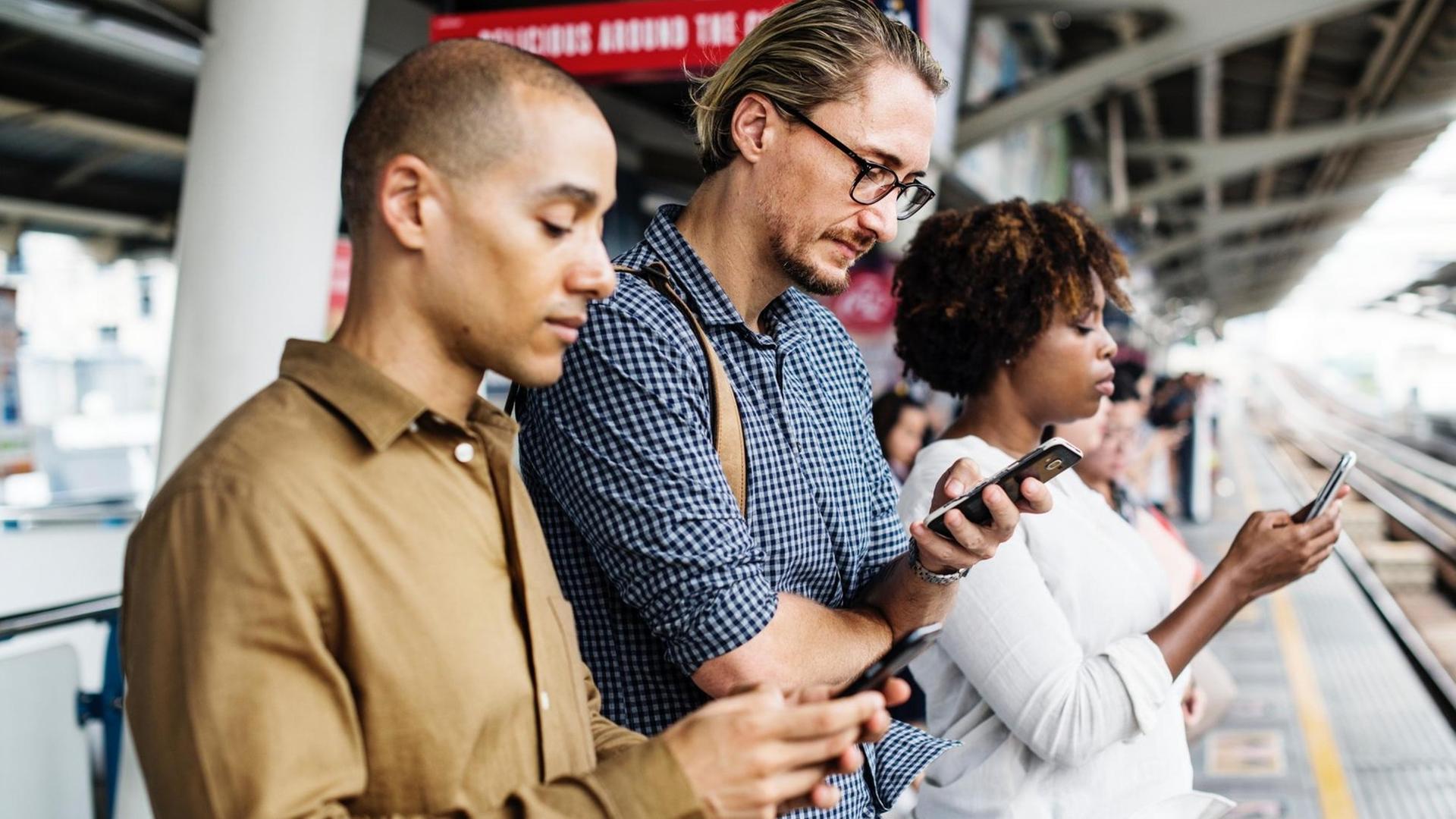 Drei Menschen stehen nebeneinander auf einem Bahnsteig und gucken auf ihre Smartphones Drei Menschen stehen nebeneinander auf einem Bahnsteig und gucken auf ihre Smartphones