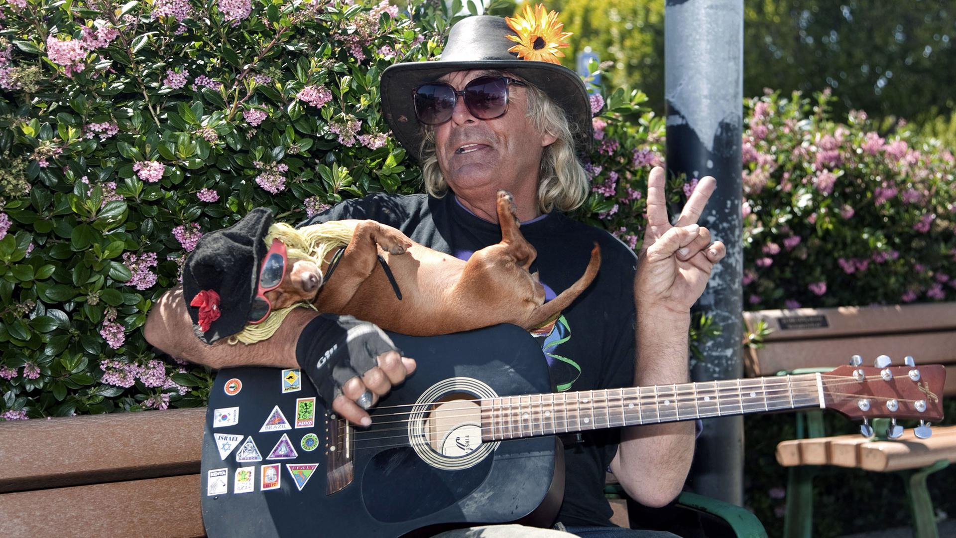 Ein älterer Hippie sitzt mit einer Gitarre und einem kleinen Hund auf dem Arm auf einer Bank und zeigt lachend das Victory-Zeichen