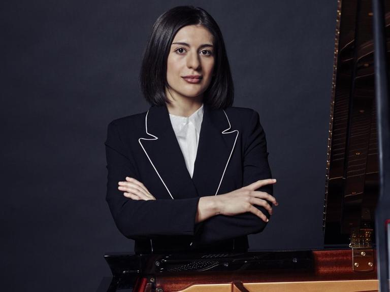 Eine junge Frau mit dunkelbraunen Haaren steht mit verschränkten Armen hinter einem Flügel. Es ist die Pianistin Mariam Batsashvili.