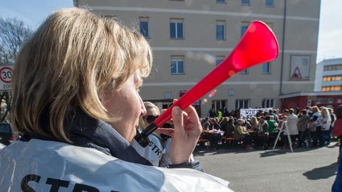Eine blonde Frau hält eine rote Tröte in der Hand und trägt einen Überwürf aus Plastik, auf dem "Streik" steht