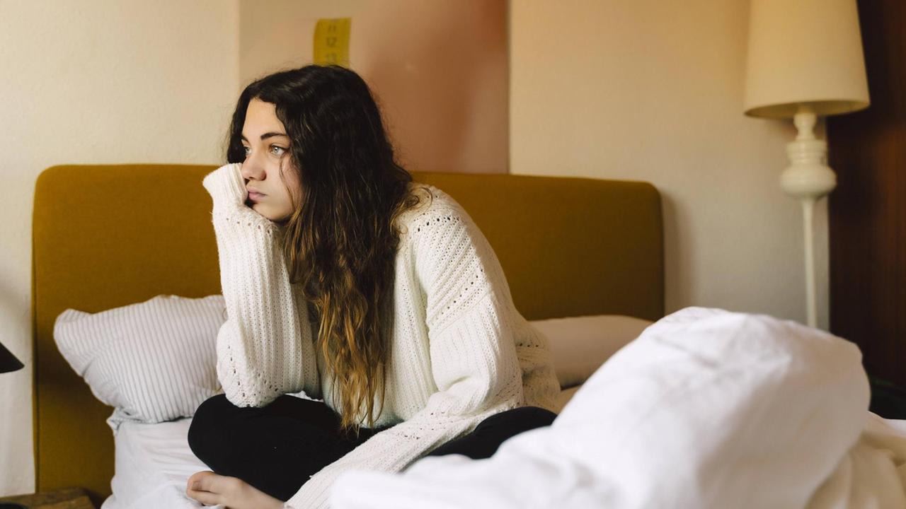 Jugendliche sitzt im Scheidersitz mit in die Hand gestützdem Kinn auf dem Bett und blickt in die Weite.