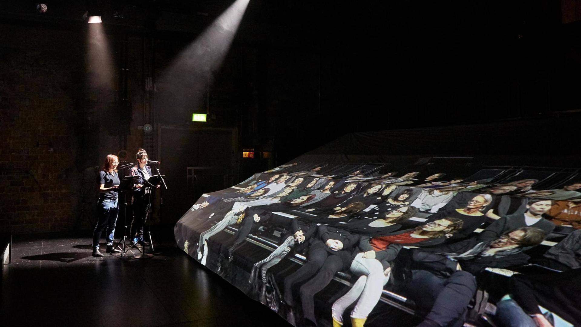 Schauspielerinnen Zeynep Bozbay und Eva Löbau stehen im Hintergrund an Mikrofonpulten im Scheinwerferlicht des dunklen Theatersaals vor mit Stoff bedeckten Sitzreihen, auf dem das Foto eines Publikums gedruckt ist. Schauspielerinnen Zeynep Bozbay und Eva Löbau stehen im Hintergrund an Mikrofonpulten im Scheinwerferlicht des dunklen Theatersaals vor mit Stoff bedeckten Sitzreihen, auf dem das Foto eines Publikums gedruckt ist.