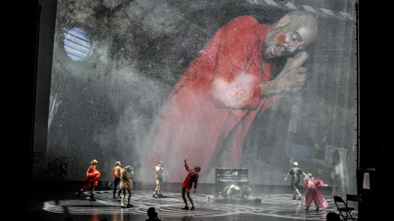 Mehrere Schauspielern auf einer Bühne stehen vor einer Leinwand-Projektion eines riesenhaft erscheinenden Menschen. Mehrere Schauspielern auf einer Bühne stehen vor einer Leinwand-Projektion eines riesenhaft erscheinenden Menschen.