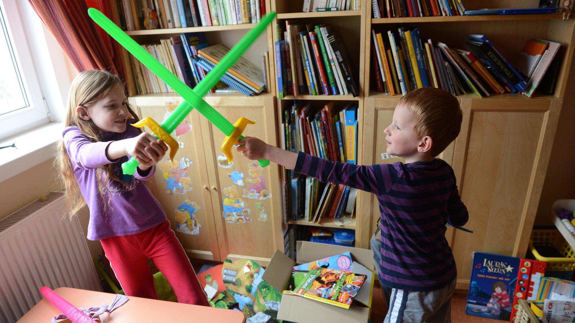 Ein achtjähriges Mädchen und ein fünfjähriger Junge "kämpfen" in ihrem Kinderzimmer mit Plastikschwertern gegeneinander. Ein achtjähriges Mädchen und ein fünfjähriger Junge "kämpfen" in ihrem Kinderzimmer mit Plastikschwertern gegeneinander.
