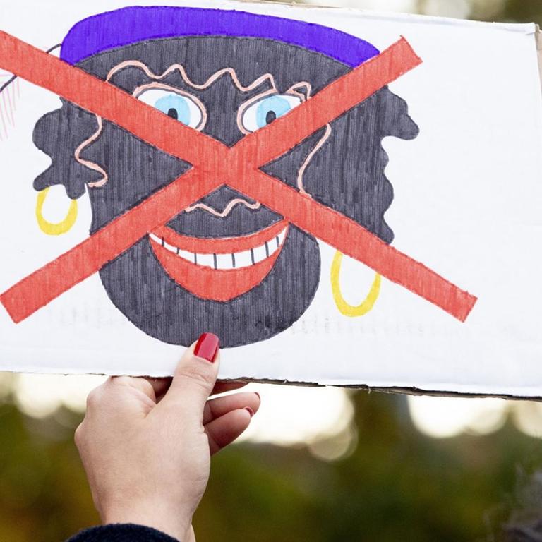 Eine Hand mit roten Fingernägeln hält ein Protestplakat, auf dem ein gezeichnetes Bild eines schwarz geschminkten "Zwarten Piet" mit goldenen Kreolen und violetter Mütze rot durchgestrichen ist. 