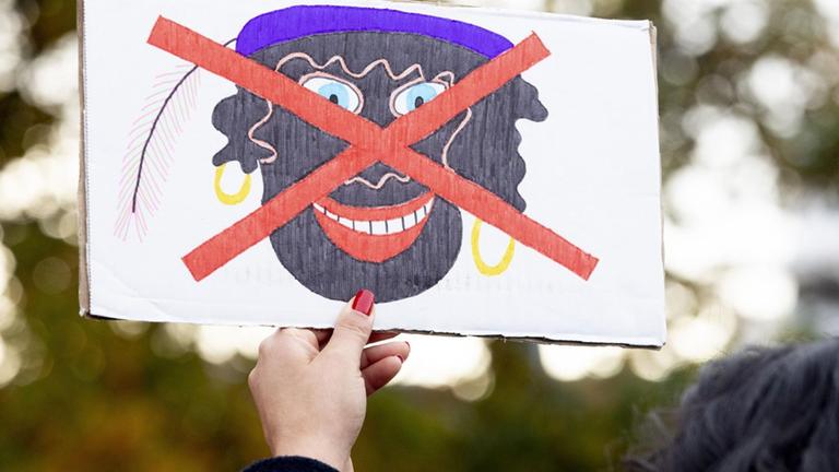 Eine Hand mit roten Fingernägeln hält ein Protestplakat, auf dem ein gezeichnetes Bild eines schwarz geschminkten "Zwarten Piet" mit goldenen Kreolen und violetter Mütze rot durchgestrichen ist. 