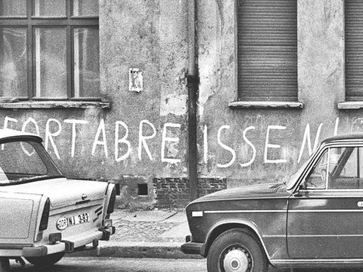 Auf einer Schwarz-weiß-Fotografie steht ein Trabant vor einer Mauer, auf der "sofort abreißen!" steht.
