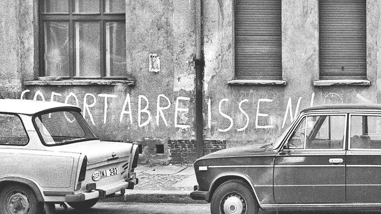 Auf einer Schwarz-weiß-Fotografie steht ein Trabant vor einer Mauer, auf der "sofort abreißen!" steht. Auf einer Schwarz-weiß-Fotografie steht ein Trabant vor einer Mauer, auf der "sofort abreißen!" steht.