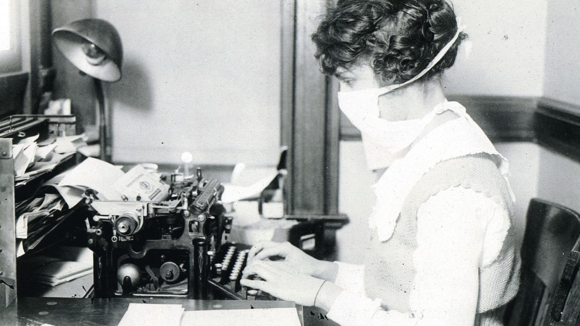 Eine Schreibkraft mit Maske an der Schreibmaschine während der Zeit der Spanischen Grippe. (1918-1920)