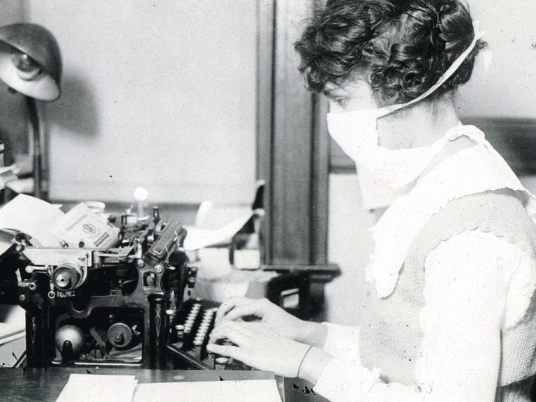 Eine Schreibkraft mit Maske an der Schreibmaschine während der Zeit der Spanischen Grippe. (1918-1920)