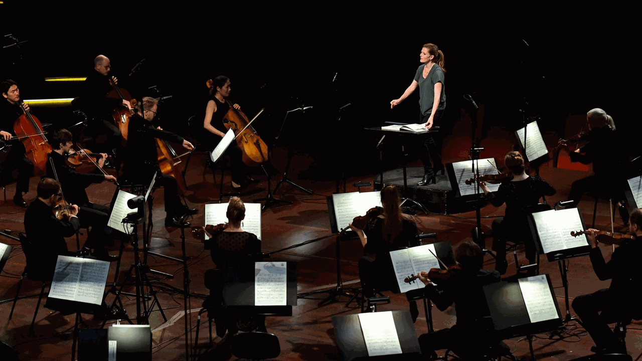 Eine Frau steht in einem grünen Shirt am Dirigentenpult und dirigiert ein Orchester, das mit relativ großen Abständen vor ihr spielt.