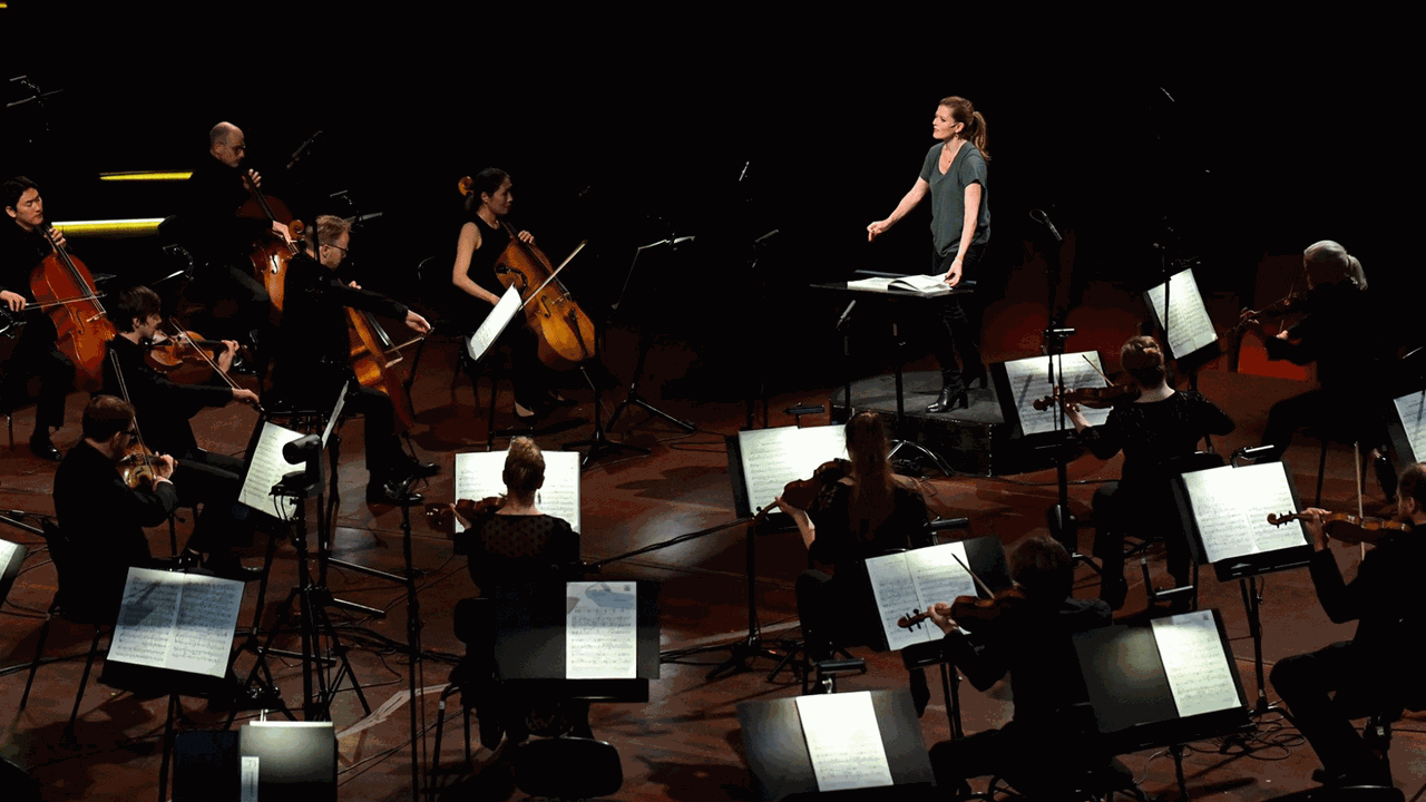 Eine Frau steht in einem grünen Shirt am Dirigentenpult und dirigiert ein Orchester, das mit relativ großen Abständen vor ihr spielt. Eine Frau steht in einem grünen Shirt am Dirigentenpult und dirigiert ein Orchester, das mit relativ großen Abständen vor ihr spielt.