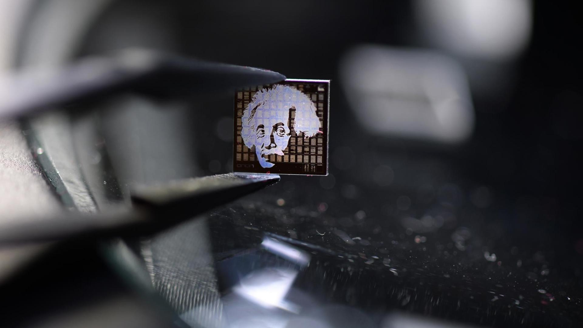 Ein Chip aus Quarzglas mit einem Albert-Einstein-Motiv darauf wird mit einer Pinzette über einen beleuchteten Mikroskoptisch gehalten. Ein Chip aus Quarzglas mit einem Albert-Einstein-Motiv darauf wird mit einer Pinzette über einen beleuchteten Mikroskoptisch gehalten.