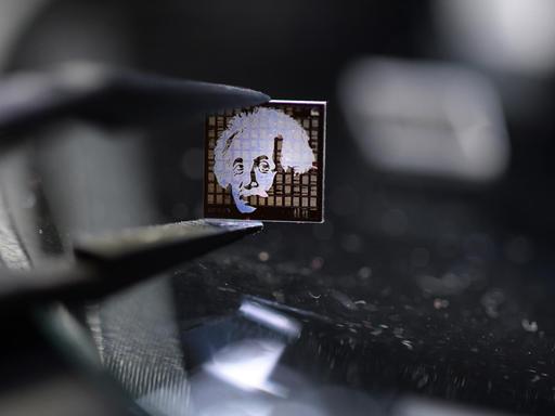 Ein Chip aus Quarzglas mit einem Albert-Einstein-Motiv darauf wird mit einer Pinzette über einen beleuchteten Mikroskoptisch gehalten.