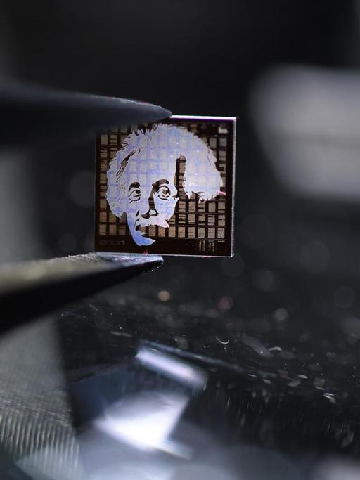 Ein Chip aus Quarzglas mit einem Albert-Einstein-Motiv darauf wird mit einer Pinzette über einen beleuchteten Mikroskoptisch gehalten.