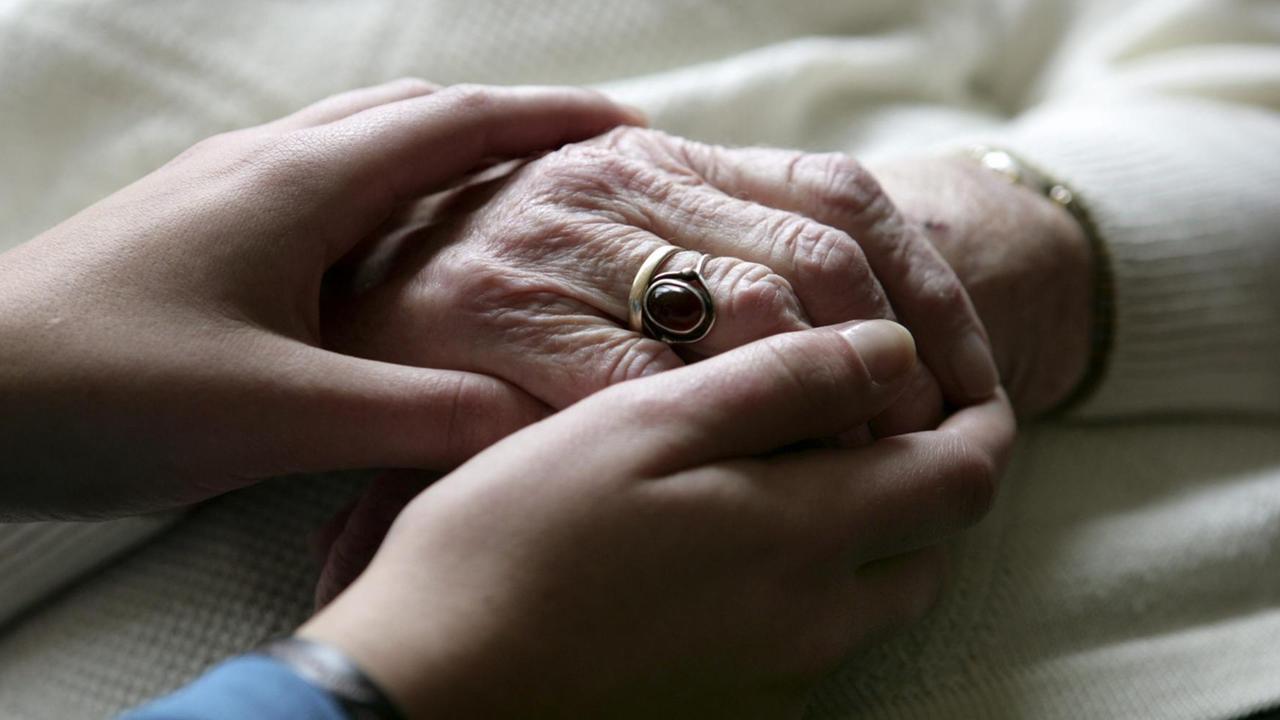  Die Hände eines jungen Menschen halten die Hände einer älteren Frau fest. Sie trägt einen Ring mit einem dunklen Stein.
