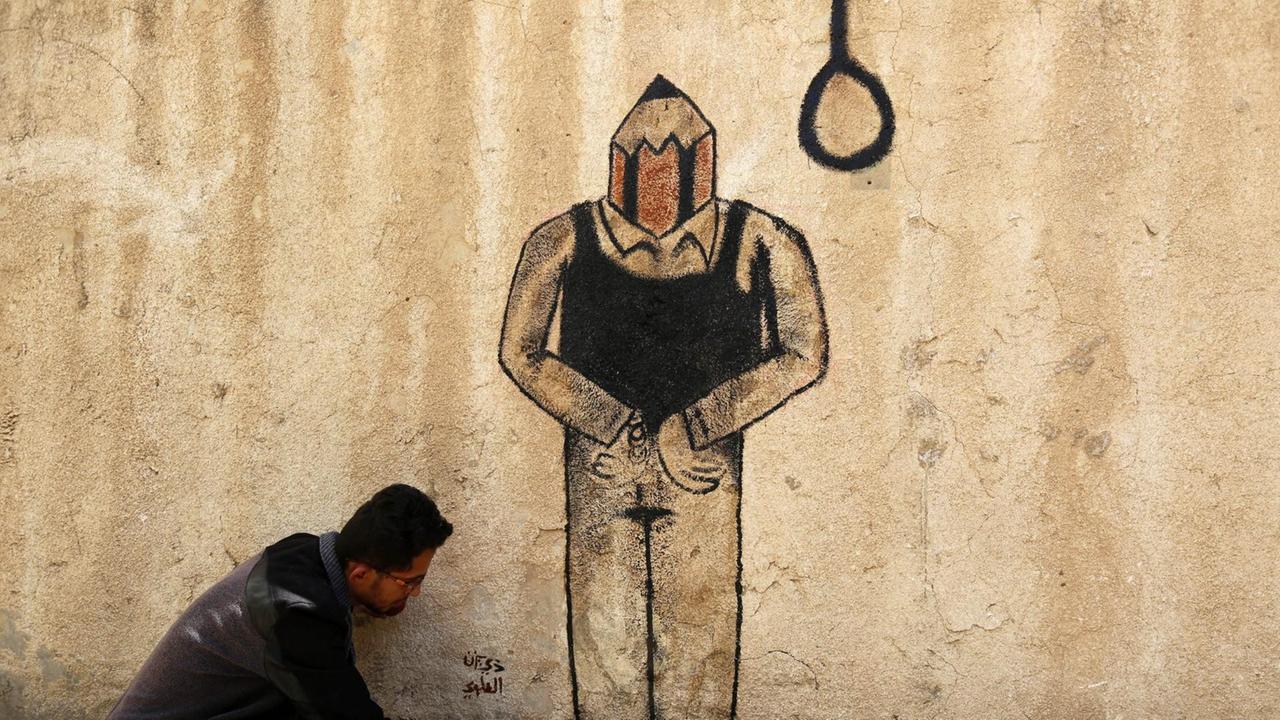 EIn Künstler malt 2015 ein Grafitti an eine Hauswand in Sanaa, das eine Person mit Bleistiftspitze anstelle des Kopfes zeigt. Neben dem Bleistiftkopf hängt ein Strick.