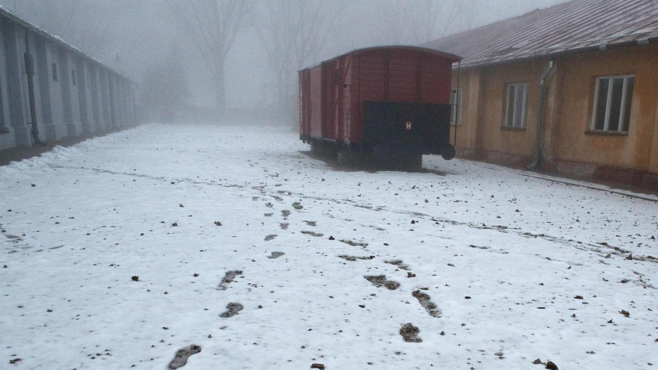 Blick auf einen verschneiten Teil des Außengeländes, wo ein Eisenbahnwagon vor Baracken steht. Blick auf einen verschneiten Teil des Außengeländes, wo ein Eisenbahnwagon vor Baracken steht.